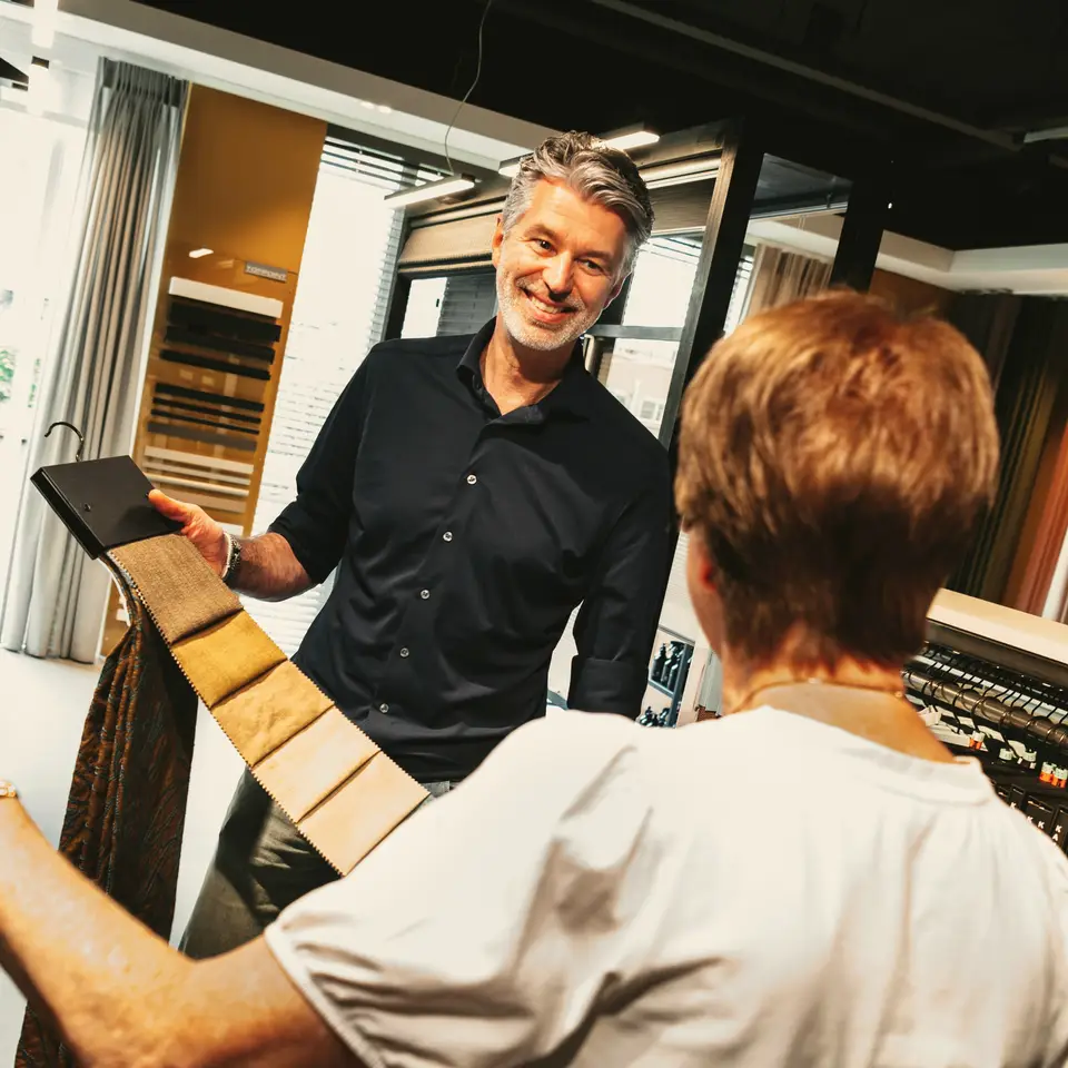 Ronald laat stalen voor gordijnen zien bij de wand met raambekleding in de showroom